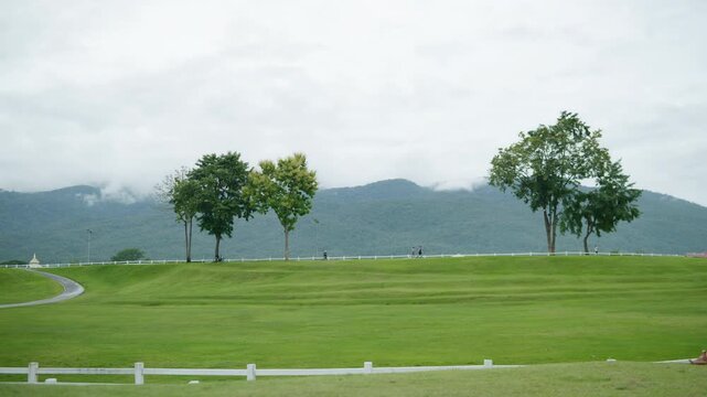 Wide view of a public park with green lawn, curving jogging track and mountain backdrop, representing healthy lifestyle, outdoor recreation, wellness and longevity concept.