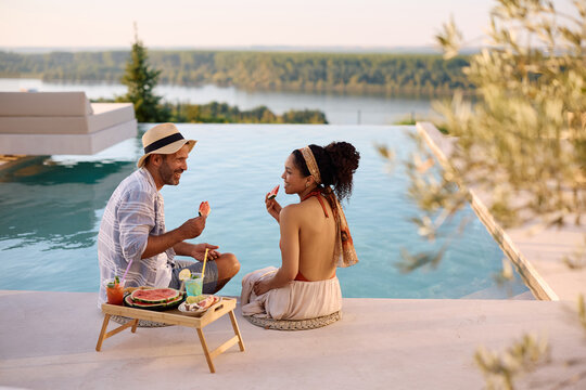 Happy multiracial couple enjoying in fresh fruit while relaxing by infinity pool on summer vacation.