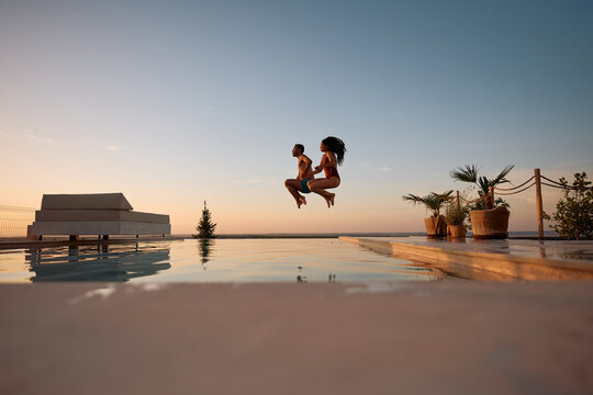 Playful couple having fun while jumping in outdoor swimming pool at sunset.