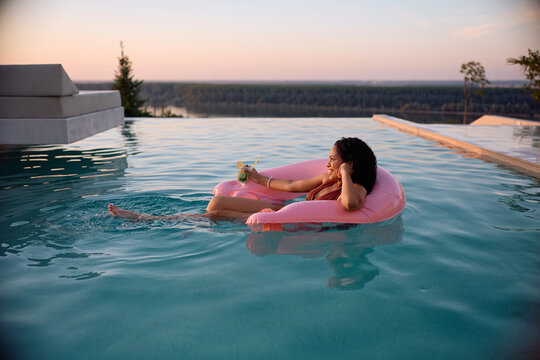 Happy black woman having a cocktail while relaxing on inflatable float in infinity pool at sunset.