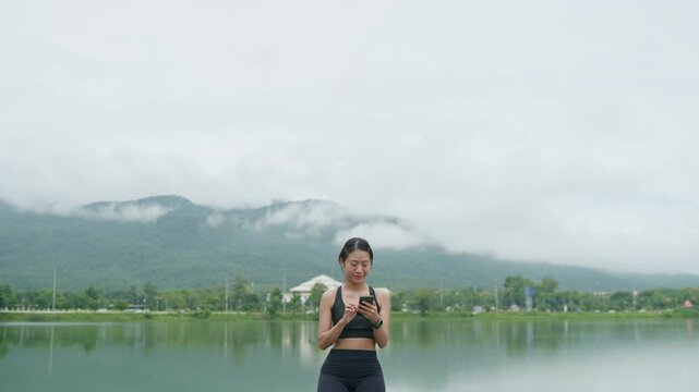 Woman using a smartphone outdoors by a scenic reservoir with mountain views, representing healthy lifestyle, wellness, active living and longevity.
