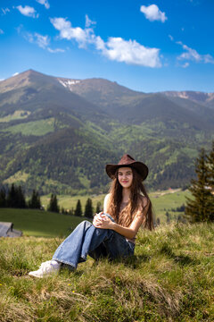 Young Woman in Cowboy Hat Sitting in Mountain Meadow. Young woman wearing a cowboy hat and jeans while sitting in a sunny meadow with forested mountains beneath a bright blue sky.