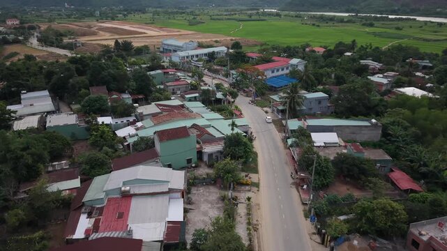 Smooth drone approach toward a peaceful rural village surrounded by lush rice fields and tropical vegetation in central Vietnam. Cinematic aerial footage showcasing traditional homes, quiet roads, and