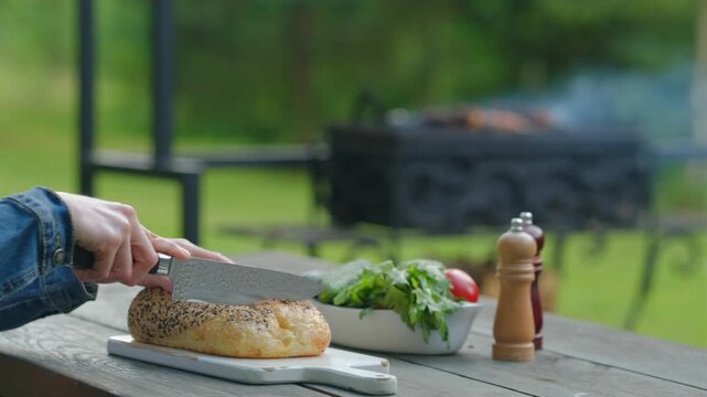 Close-up view of hands slicing fresh bread in backyard with barbecue in background