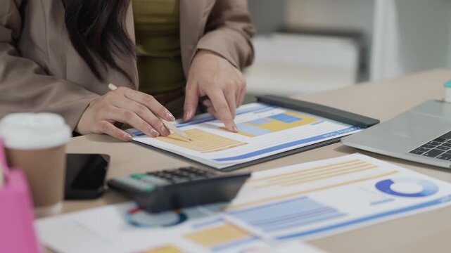 A smiling Asian businesswoman is working with a laptop and financial documents at her desk in the office.