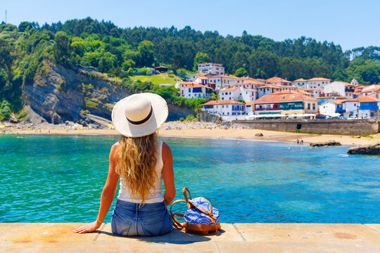 Woman tourist with straw hat sitting by turquoise ocean looking at traditional coastal village in Asturias, tour tourism in Spain. Tazones