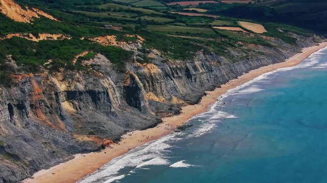 Drone view along dramatic layered cliffs on the Dorset Jurassic Coast, with sandy beach, turquoise sea, waves and countryside above the cliff edge. Scenic British coastal landscape useful for travel