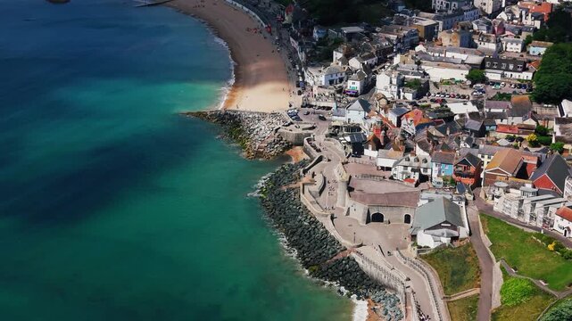 Drone view over Lyme Regis on the Dorset Jurassic Coast, showing the Cobb harbour wall, seafront, sandy beach, turquoise water and coastal town on a sunny summer day.
