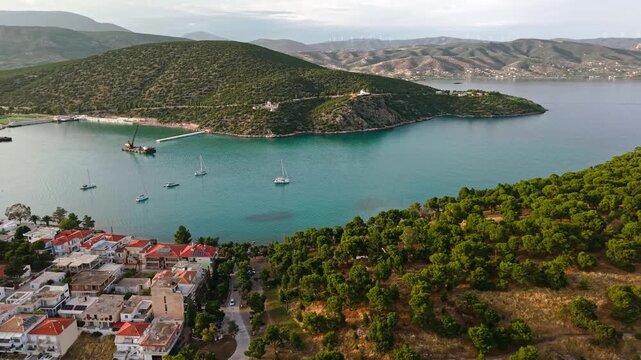 Wide drone shot of Ermioni, Greece at golden hour. Lush green pine forest bordering a calm turquoise sea bay with anchored yachts and mountains