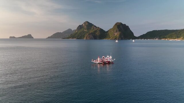 Beautiful high-angle drone shot of traditional paraw sailboats with striped sails cruising on calm sea water near dramatic limestone mountain islands under a soft sunset sky in the Philippines.