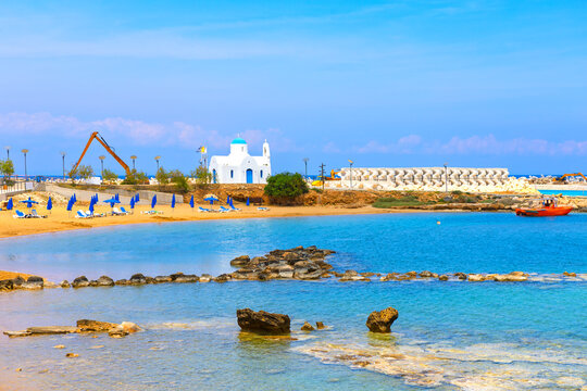 Louma Beach in Protaras from Cyprus features traditional white and blue church overlooking a scenic bay. Sun loungers sit on golden sand beside turquoise water under a clear summer sky