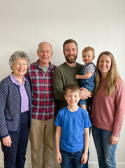 Obraz premium Multigenerational family portrait showing grandparents parents and two children smiling together against a simple white background in bright indoor lighting