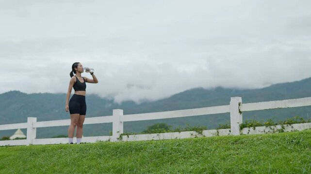 Wide shot of a female runner drinking water beside a lake with mountain scenery, representing outdoor fitness, hydration, wellness, active lifestyle and longevity.