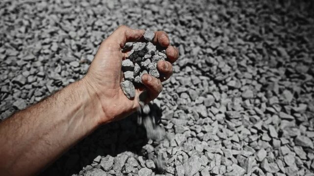 Man inspecting quality of crushed stone pile with repeated hand sampling under bright sunlight, showing rough aggregate texture, industrial setting, low ground viewpoint, strong contrast