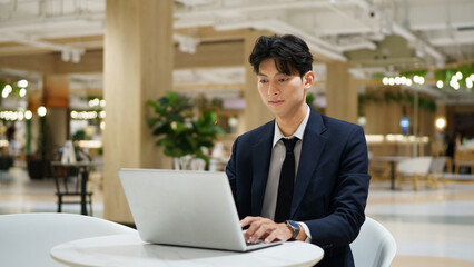 Fototapeta premium Young businessman in navy suit smiling while contemplating new strategies and opportunities, working on a laptop in a contemporary co-working space