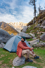 Fototapeta premium Man sitting on a rock near a camping tent at Kareri Lake in the mountains of Himachal Pradesh
