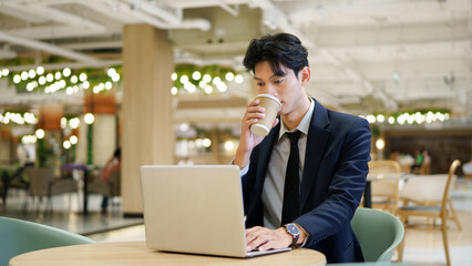 Fototapeta premium Young businessman in navy suit holding coffee cup while contemplating new strategies and opportunities, working on a laptop in a contemporary co-working space
