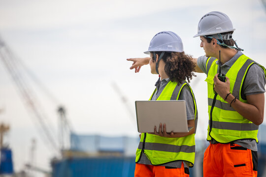 Engineers in safety gear and hard hats pointing and discussing project at shipping port, Construction managers planning and reviewing site progress at industrial facility with cranes