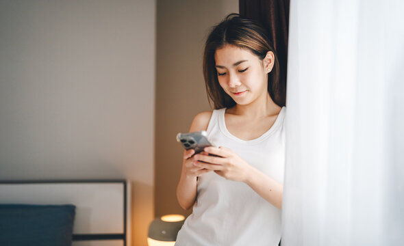 Young adult asian woman texting phone for telemedicine, digital social network dating app, after wake up. Bedroom background with curtain. People using AI technology stay home lonely