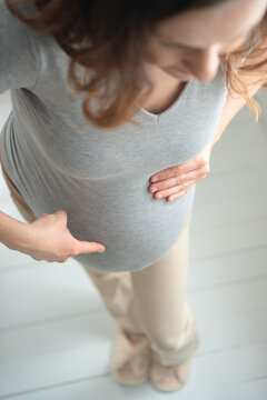 A pregnant woman points at her belly from above, announcing her bump with joyful excitement. The expectant mother presenting a pregnancy sign radiates a cheerful, proud moment in a clean studio.