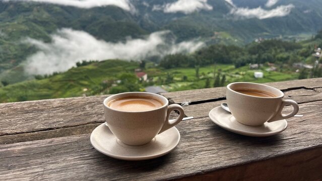 Two cups of hot coffee on a wooden table with a scenic mountain view. Morning coffee for two on a rustic terrace overlooking foggy green valley and mountains in Sapa,  Vietnam