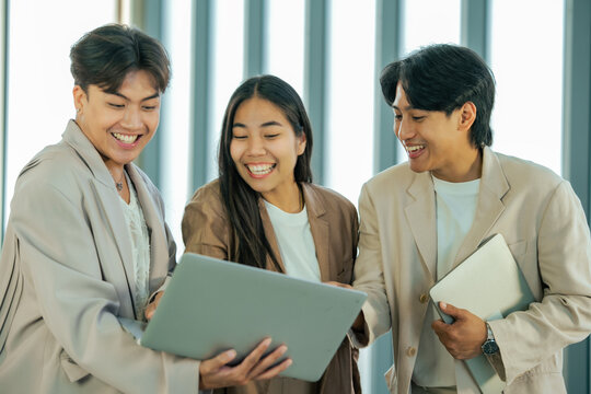 Smiling Asian business professionals collaborate around laptop in modern office. Creative teamwork, project planning, knowledge sharing, technology, employee engagement, business communication.