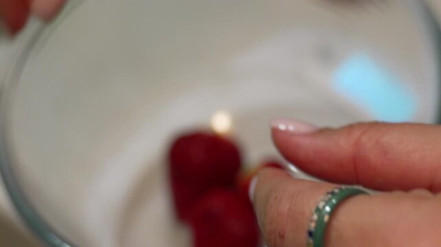 Fresh strawberry hulling process. Close-up high-angle shot showcasing a woman removing green stems from organic fruit before placing them into a glass bowl.