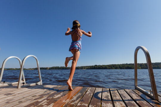 A courageous child with goggles makes a confident leap from a wooden dock into deep blue lake water demonstrating bravery and personal growth through an adventurous outdoor challenge on a sunny day