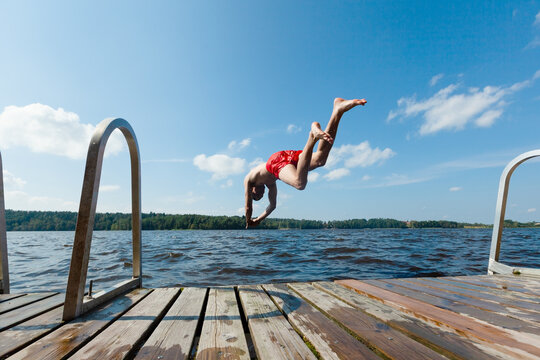 Man dives off dock with arms spread knowing how to fall safely into lake water. Guy in red trunks shows proper entry technique midair on summer day. Skilled diver demonstrates safe plunge form