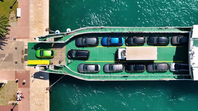 Ferry is at dock. Vehicles are lined up for boarding. Passengers are disembarking and boarding the ferry. Water is visible around the ferry.