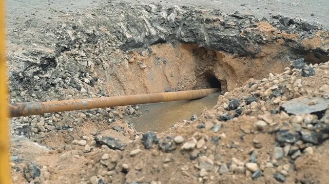 construction on a road where a trench has been dug to install a pipe. The location is marked by safety barriers and exposed soil amidst ongoing utility work