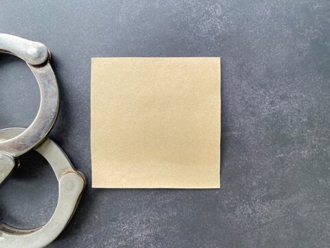 Blank sticky note beside metal handcuffs on a dark background for law enforcement, crime prevention and legal awareness concepts.