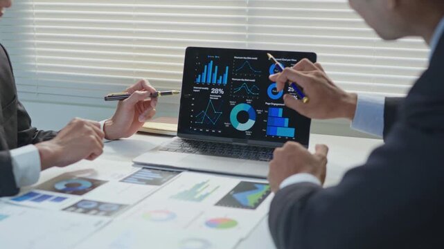 Business professionals discussing and explaining data charts on a laptop screen during an office meeting, with reports and documents on the desk, representing teamwork, analysis and strategic planning