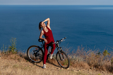 Fototapeta premium Cyclist woman nature, athletic female wearing red sportswear posing with bicycle on a cliff above the blue sea at sunny day