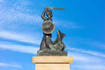 This bronze sculpture, an iconic symbol of Warsaw, depicts a mythical mermaid wielding a sword and shield against a vibrant, cloud-streaked cerulean sky. © Anton Kustsinski