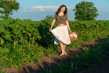 Fototapeta premium Young woman in white skirt walking through sunflower field with wicker basket