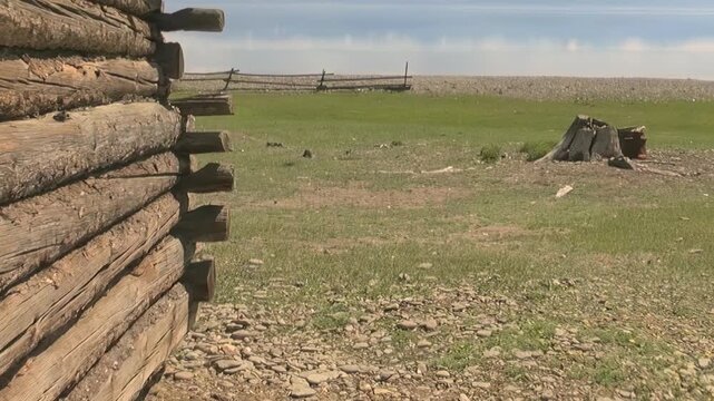 Rustic log structure frames sprawling green field in Mongolia, revealing distant wooden fence and old tree stump. This expansive natural beauty thrives near Lake Khuvsgul on bright, peaceful day.