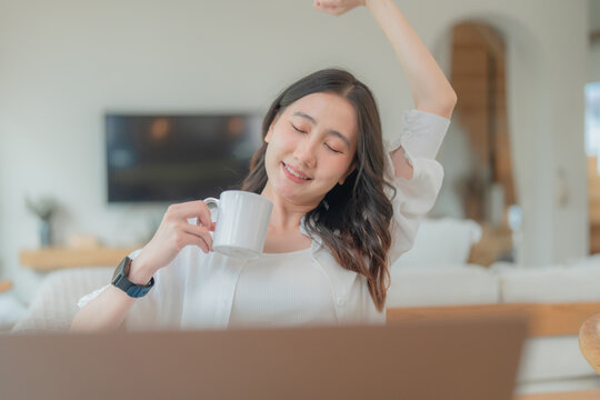 Young Asian woman raising arm to stretch and relax muscles with hot drink during a break from computer work in a bright living room.