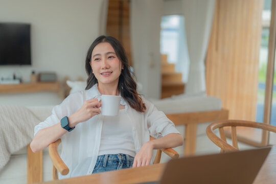 Young Asian woman relaxing with a hot drink during remote work, sitting at wooden table with laptop in a bright modern home interior.