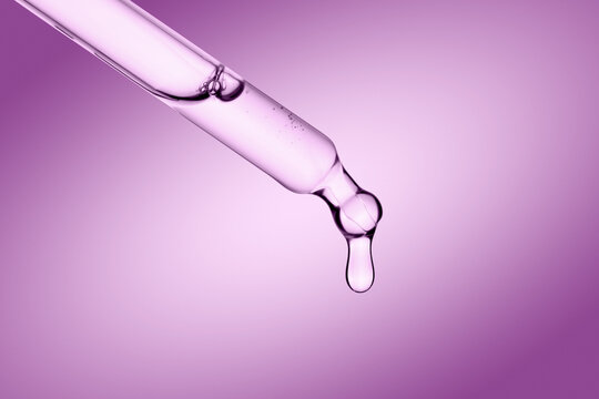 Dropper with a clear drop of cosmetic serum or gel falling on pink background.