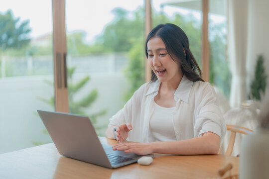 Young Asian woman working on computer beside glass door with green nature outside, cheerful freelancer at bright home workspace.