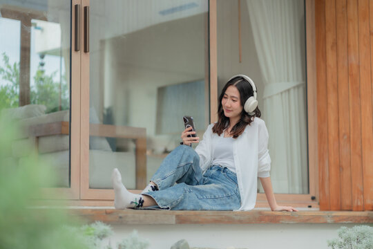 Young Asian woman reading social media and chatting on mobile phone outside glass door, relaxing on home deck in casual jeans.