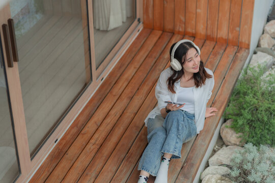 Young Asian woman listening to music from mobile phone on house deck, sitting by rock garden, enjoying leisure audio entertainment.