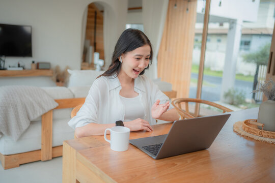 Young Asian woman greeting family on online video call at home, casual conversation with friends through computer with coffee mug.