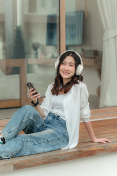 Young Asian woman listening to podcast on smartphone smiling at camera, vertical lifestyle photo on wooden deck of modern house.