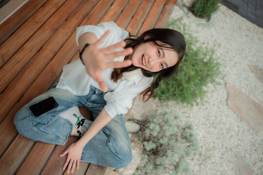 Cheerful young Asian woman with headphones around neck smiling and stretching palm to camera, fun candid moment on house terrace.
