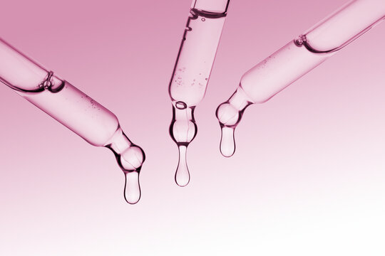 Dropper with a clear drop of cosmetic serum or gel falling on pink background.
