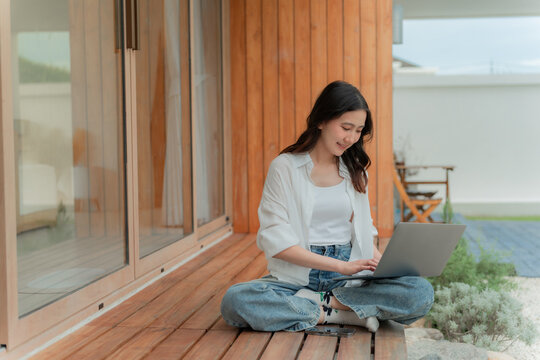 Young Asian woman balancing work and relaxation outdoors, computer on lap at house patio with garden, comfortable green workspace.