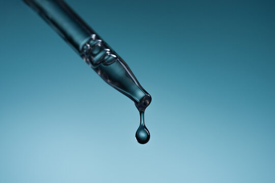 Dropper with a drop of cosmetic serum or gel falling on blue background.