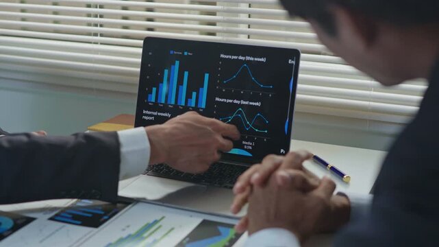 Business professionals discussing and explaining data charts on a laptop screen during an office meeting, with reports and documents on the desk, representing teamwork, analysis and strategic planning